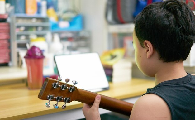 Niño de Asia aprende a tocar la guitarra en línea por medio de una tableta en la vista posterior de la casa de la habitación