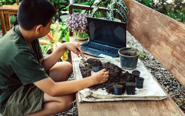 Niño asiático aprende a cultivar flores en macetas a través de la enseñanza en línea de palear tierra en macetas