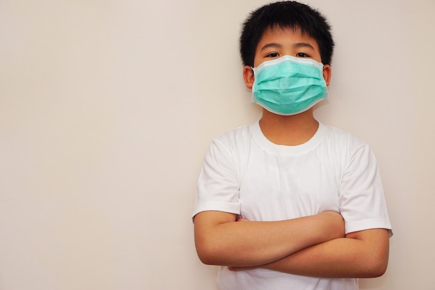 Un niño asiático con una camiseta blanca cruza el brazo mirando directamente a la cámara, con máscara médica en la cara y ojos felices. Concepto de salud, tratamiento, protección, resistencia y lucha.