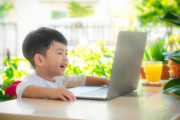 Foto niño asiático divertido y viendo dibujos animados video en él computadora cuaderno en la mesa en el jardín de la casa