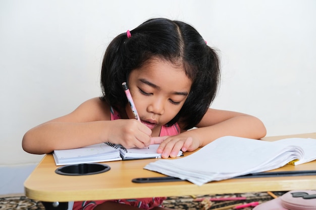 Niño asiático escribe algo en el libro con lápiz sobre la mesa de estudio