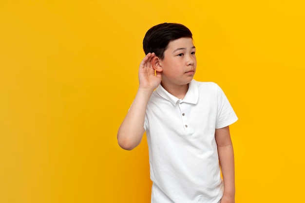 niño asiático escuchando a escondidas sobre fondo amarillo niño coreano sostiene la mano cerca de la oreja y escucha