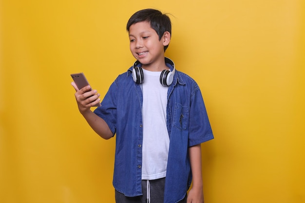 Niño asiático feliz en estilo denim casual mirando el teléfono inteligente aislado sobre fondo amarillo