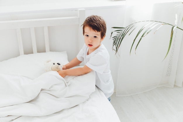 Un niño asiático feliz limpia su habitación y dobla una manta en el dormitorio de su casa. El niño hace la cama y ajusta la manta en la cama grande.