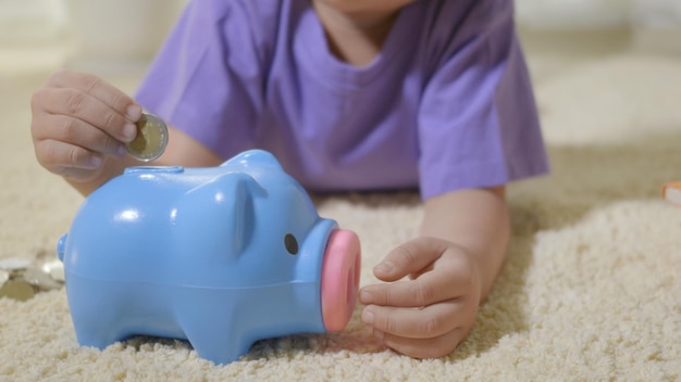 Foto un niño asiático feliz de preescolar poniendo monedas de dinero en la ranura del cerdo de cara azul.