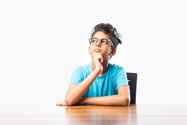 Niño asiático indio o niño sentado en una mesa o escritorio, mirando a cemara, pensando o señalando algo