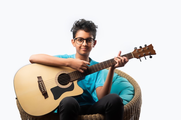 Niño asiático indio tocando la guitarra acústica mientras está sentado contra la pared blanca