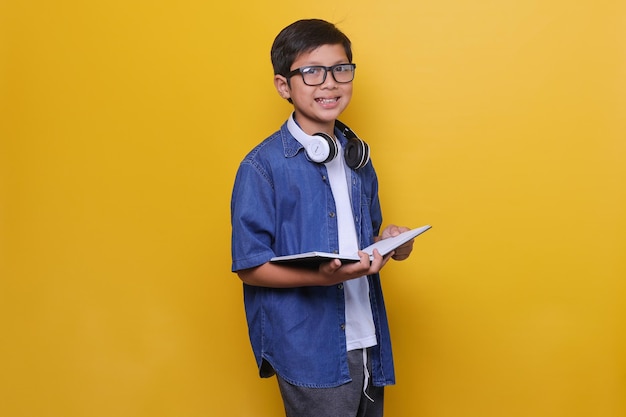 Niño asiático inteligente con ropa informal, auriculares y anteojos sosteniendo un libro y sonriendo al