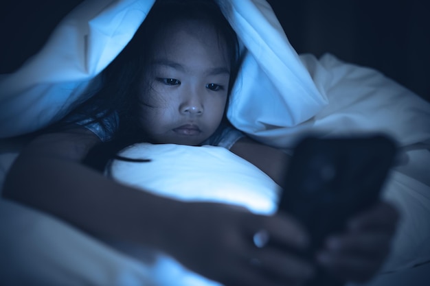 Niño asiático jugando en el teléfono inteligente en la cama por la noche La chica Addict social media