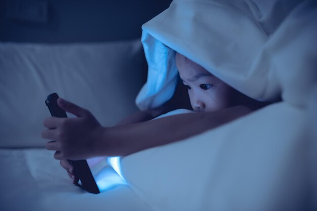 Niño asiático jugando en el teléfono inteligente en la cama por la noche La chica Addict social media