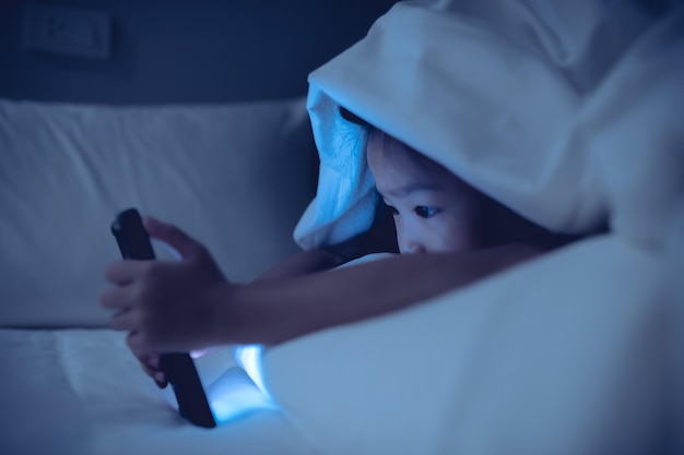 Niño asiático jugando en el teléfono inteligente en la cama por la noche La chica Addict social media