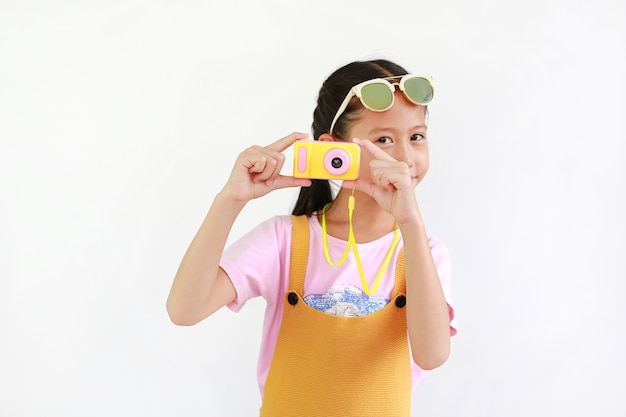 Niño asiático lindo de la niña que toma el cuadro con el juguete de la cámara de fotos aislado en el fondo blanco.