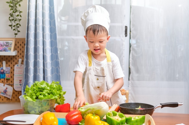 Niño asiático lindo del niño pequeño que lleva el sombrero y el delantal del cocinero que se divierten preparando, cocinando la comida sana en cocina