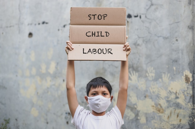 Un niño asiático con una máscara médica que levanta un letrero dice "Alto al trabajo infantil cuando la campaña"