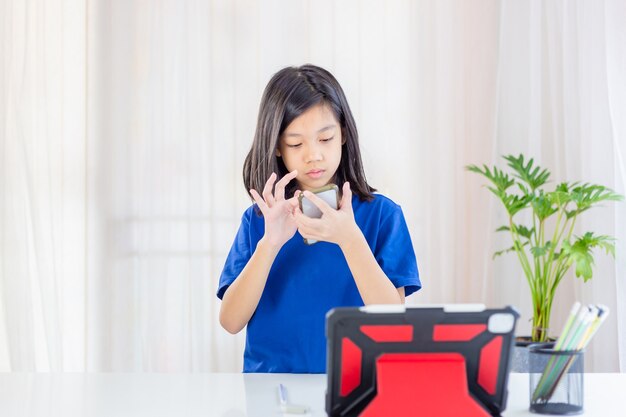 Niño asiático de pie y usando un teléfono inteligente con una tableta y lápices en la mesa de la sala de estar