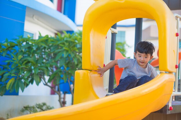 Niño asiático que juega la diapositiva en el patio bajo la luz del sol en verano, niño feliz en guardería o patio de la escuela preescolar.