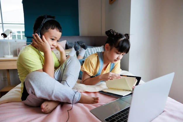 Niño asiático que usa una computadora portátil y auriculares para estudiar en línea con un maestro de videollamadas y que una linda hermana lo ayude en el dormitorio