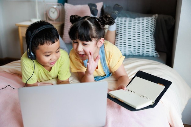 Niño asiático que usa una computadora portátil y auriculares para estudiar en línea con un maestro de videollamadas y que una linda hermana lo ayude en el dormitorio