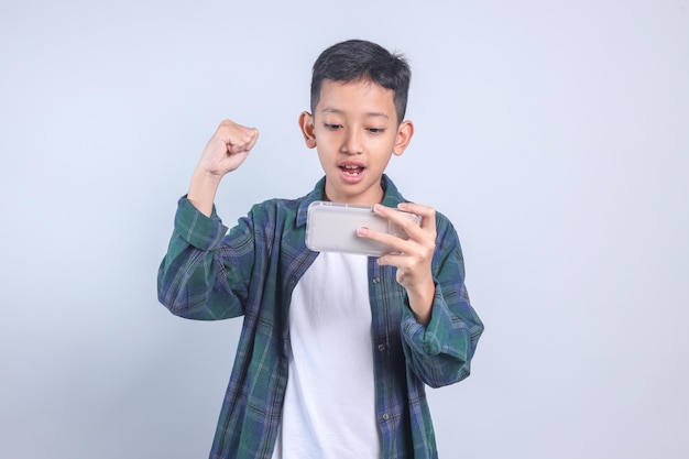 Niño asiático en ropa casual sosteniendo un teléfono inteligente mientras hace el gesto ganador celebrando la victoria del éxito