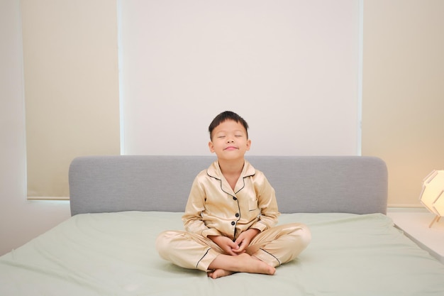 Niño asiático sentado en la cama y practicando meditación por la mañana después de despertarse en el dormitorio