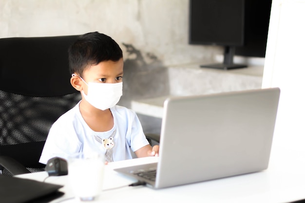 Niño asiático sentado en la mesa con el portátil y la mascarilla médica y preparándose para la escuela. Concepto de educación en línea. Estudio de lección de clase de conferencia de video llamada en línea.