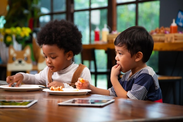 Un niño asiático sonriente comiendo comida con un amigo afroamericano en una fiesta de cumpleaños en casa. Felicidad, familia, gente y fiesta de cumpleaños.