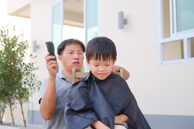 Foto un niño asiático sonriente y lindo que se corta el pelo en el patio trasero de su casa el padre hace un corte de pelo para su hijo durante el encierro corte de pelo en casa mientras está en aislamiento de cuarentena durante la crisis de salud de covid19