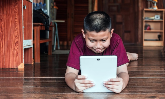 Un niño asiático sonriente sentado en el suelo de madera y jugando en una tableta digital en casa