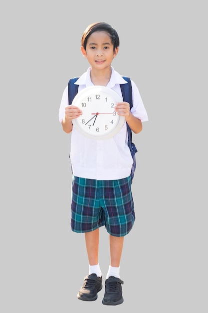 Un niño asiático en uniforme de escuela está de pie sosteniendo un reloj blanco y sonriendo gris aislado