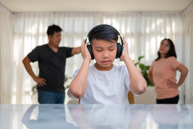 Niño asiático usando auriculares y tocando música a todo volumen para no escuchar peleas mientras los padres