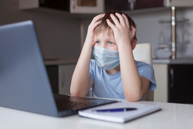 Niño asustado en máscara médica stydying en casa, haciendo la tarea escolar