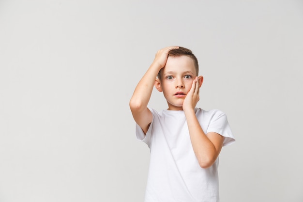 Niño aterrorizado en camiseta blanca con ambas manos en la cabeza sobre fondo gris