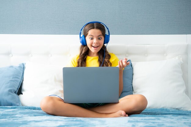 Niño con auriculares, relájese en la cama en casa usando una computadora portátil Niño con auriculares, navegue por Internet en la computadora