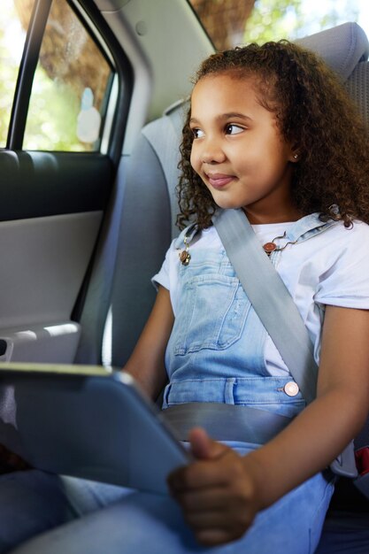 Foto niño en automóvil sonriendo y tableta en viaje por carretera con cinturón de seguridad por seguridad y dispositivo para ver videos educativos o juegos en línea tecnología internet y viaje niña feliz en el asiento trasero para conducir o compartir automóvil