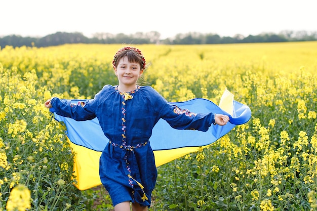 Niño con bandera ucraniana en campo de colza Una niña con una camisa bordada corre por el campo con la bandera ucraniana en sus manos