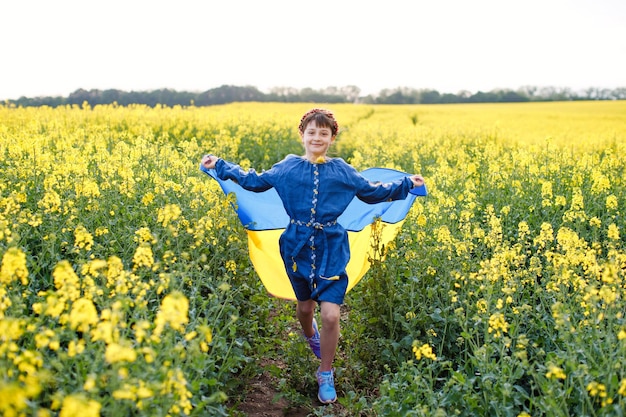 Niño con bandera ucraniana en campo de colza Una niña con una camisa bordada corre por el campo con la bandera ucraniana en sus manos