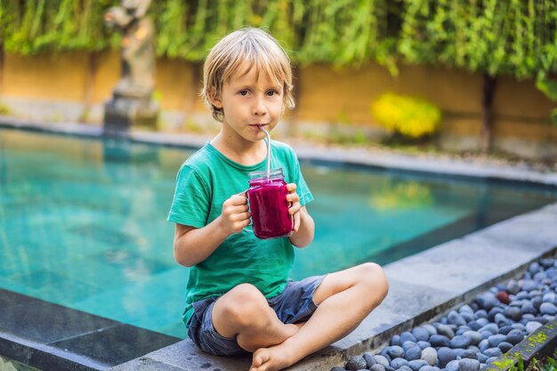 Niño bebe batido de fruta de dragón con pajita de acero junto a la piscina