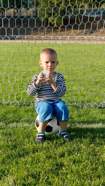 Niño bebiendo jugo de fruta refrescante mientras toma un descanso sentado en la pelota en las porterías mientras juega al fútbol