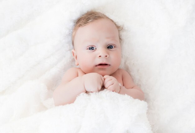 Un niño blanco de cuatro meses con ojos azules yace sobre una manta blanca y se cubre con él