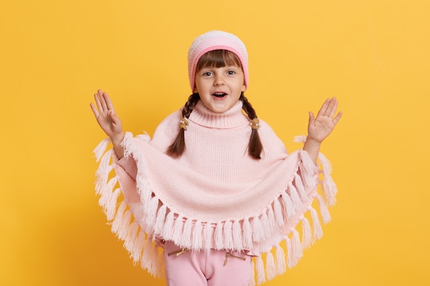 Niño con la boca abierta extendiendo las manos con entusiasmo, mira al frente, vistiendo gorra, poncho de punto y pantalones