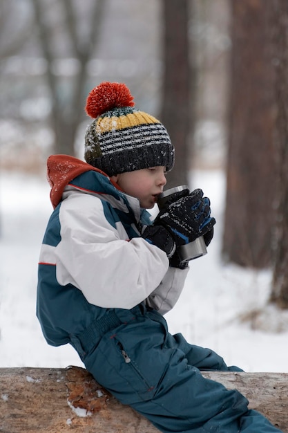 Niño en el bosque de invierno bebe té caliente de un termo. Retrato de niño con sombrero de punto en bosque nevado.