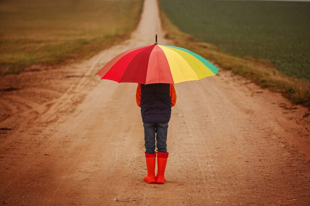 Niño en botas de goma naranja con coloridos paraguas bajo la lluvia en otoño. vista trasera