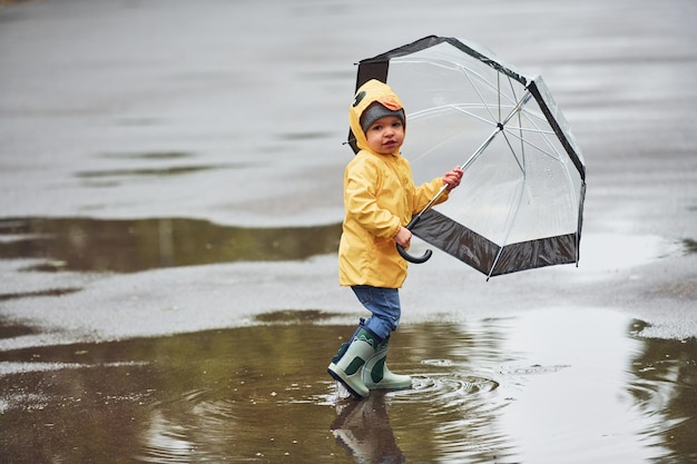 Niño con botas impermeables amarillas y paraguas jugando al aire libre después de la lluvia