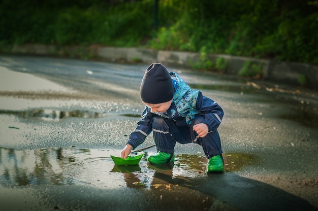 Niño en botas de lluvia verde jugando con un barco de papel verde en un charco