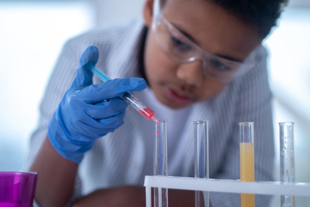 Un niño de cabello oscuro con una bata de laboratorio sosteniendo una jeringa con un reactivo y luciendo ocupado