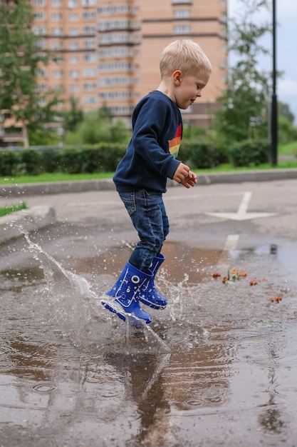 Un niño con cabello rubio en botas de goma azul salta a un charco