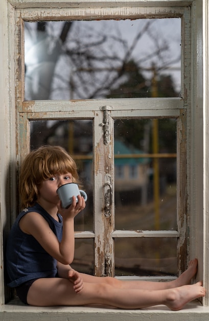 Un niño con cabello rubio se sienta en un viejo alféizar en mal estado y bebe de una taza de hierro