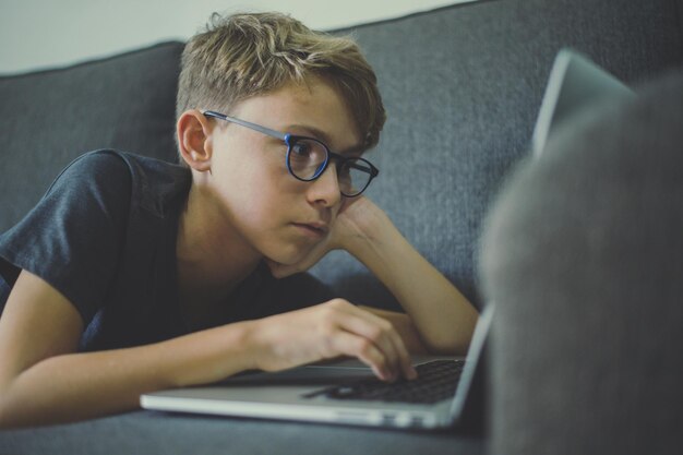 Niño de cabello rubio usando una computadora portátil mientras está acostado en el sofá en casa