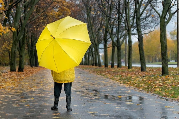 Foto niño caminando con un gran paraguas amarillo en el parque lluvioso de otoño fondo de hojas caídas vista posterior