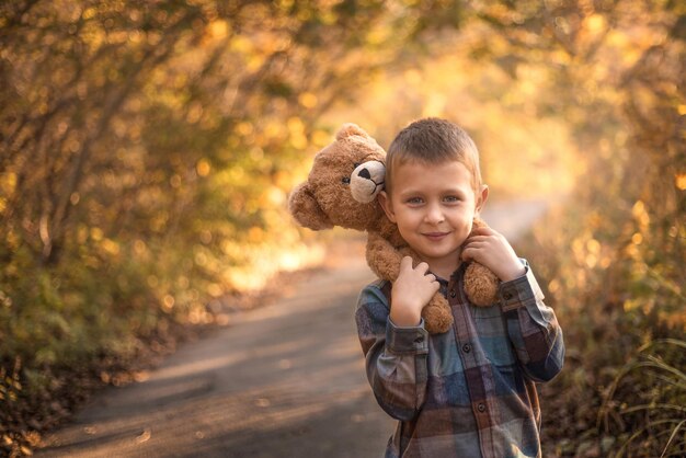 un niño se para en un camino rural en el bosque sobre sus hombros se sienta un oso de juguete